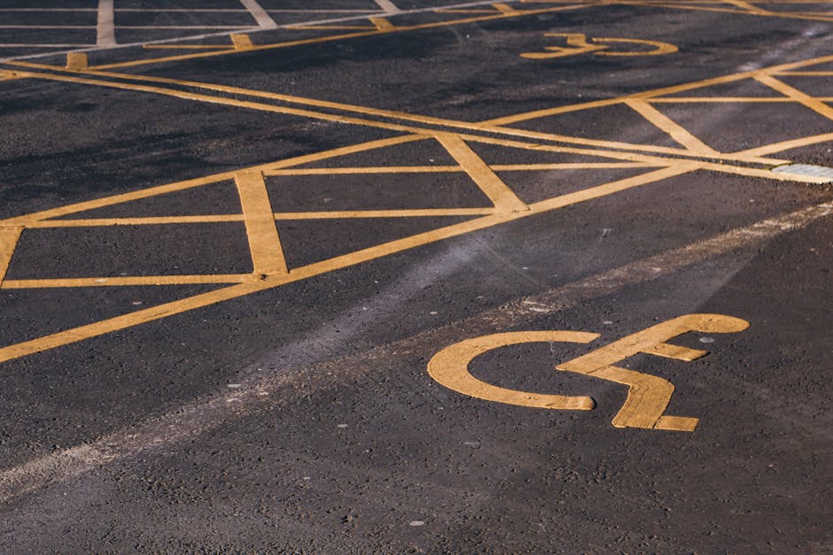 A close-up view of a parking lot surface with yellow-painted lines marking accessible parking spaces and a prominent yellow disabled parking symbol on the asphalt. The parking area is outdoors with no vehicles present, and the surface shows signs of wear and some minor dirt. The painted lines and symbol are clearly visible and well-defined. This image highlights the importance of accessible parking arrangements in residential or commercial property relocations, which are part of the logistics managed by Man with Van Bickley during home removals or furniture transport services, ensuring proper access during the loading and unloading process at a property on the specified page titled Avoid move-day delays in Bickley: parking & access fixes, BICKLEY.