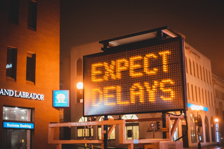 A brightly lit digital roadside sign displays the message 'EXPECT DELAYS' in orange LED lights, mounted on a portable frame with a white support structure. Behind the sign, there is a background of urban buildings with illuminated windows and streetlights, indicating evening or night-time conditions. The scene is set on a city street, and the sign appears to be part of logistical or traffic management measures related to home relocation or moving services, suitable for describing transportation or access challenges during a house move by Man with Van Bickley.