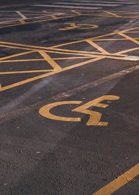 A close-up view of a parking lot surface with yellow-painted lines marking accessible parking spaces and a prominent yellow disabled parking symbol on the asphalt. The parking area is outdoors with no vehicles present, and the surface shows signs of wear and some minor dirt. The painted lines and symbol are clearly visible and well-defined. This image highlights the importance of accessible parking arrangements in residential or commercial property relocations, which are part of the logistics managed by Man with Van Bickley during home removals or furniture transport services, ensuring proper access during the loading and unloading process at a property on the specified page titled Avoid move-day delays in Bickley: parking & access fixes, BICKLEY.
