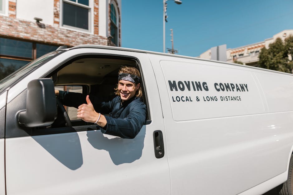 A young man with long, curly hair and a headband, smiling and giving a thumbs-up gesture while sitting in the driver's seat of a white moving van. The van has branding indicating it is operated by a removal company offering local and long-distance furniture transport services, with the text 'MOVING COMPANY, LOCAL & LONG DISTANCE' visible on the side. The van is parked outside a residential building with a brick facade and large windows, under a clear blue sky. Behind the vehicle, there are buildings, utility poles, and some trees, suggesting an urban or suburban environment suitable for house removals and relocation logistics. The scene is well-lit with natural daylight, emphasizing the vehicle’s readiness for home relocation or furniture transport involving packing and loading processes.