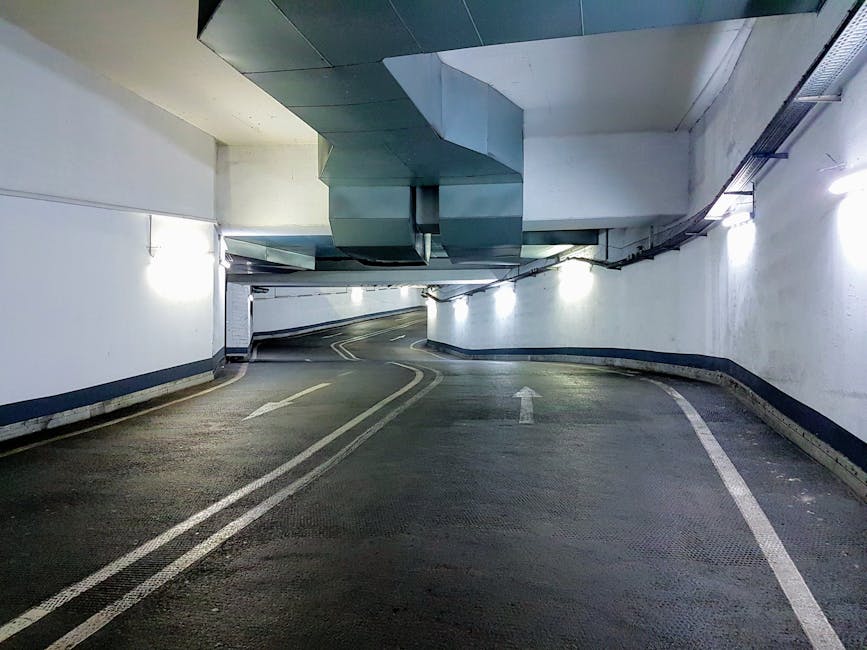 Inside a narrow underground parking garage with a curved road surface marked by white directional arrows and lane lines, illuminated by overhead fluorescent lights. The walls are painted white with a dark blue trim along the lower edge, and the ceiling features large metallic ductwork and ventilation systems. The floor is textured black asphalt with visible minor wear. This setting resembles the driveway or loading area where a home relocation might occur, with no vehicles or objects present. The environment supports furniture transport and packing and moving activities typically associated with house removals, as transported items would be loaded or unloaded in this space. The overall scene is clean and organized, indicating a professional moving process, possibly involving company Man with Van Bickley.