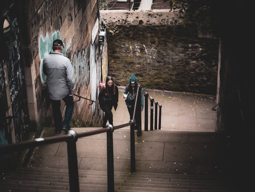 Inside a narrow underground parking garage with a curved road surface marked by white directional arrows and lane lines, illuminated by overhead fluorescent lights. The walls are painted white with a dark blue trim along the lower edge, and the ceiling features large metallic ductwork and ventilation systems. The floor is textured black asphalt with visible minor wear. This setting resembles the driveway or loading area where a home relocation might occur, with no vehicles or objects present. The environment supports furniture transport and packing and moving activities typically associated with house removals, as transported items would be loaded or unloaded in this space. The overall scene is clean and organized, indicating a professional moving process, possibly involving company Man with Van Bickley.