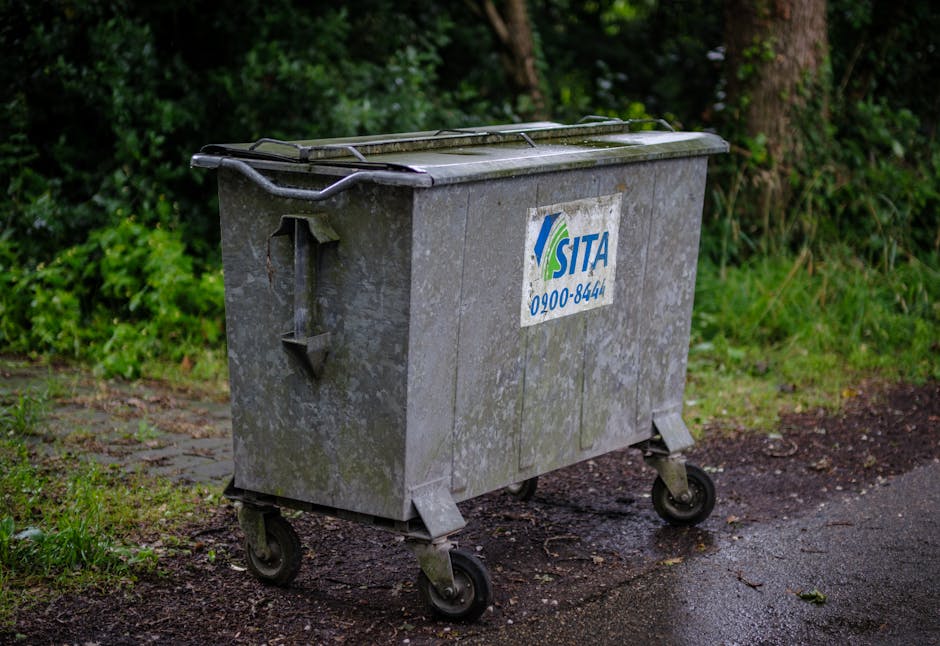 A large, weathered metal waste bin with a lid, situated on a paved driveway next to a grassy and leafy area with dense green foliage in the background. The bin has four black caster wheels and a white sticker with blue and green text displaying the SITA logo and contact number. The bin appears to be used for general waste disposal, with some signs of dirt and moss on its surface. Its position suggests it is outside a property, possibly awaiting collection, as part of the home relocation process or the clearing of waste during packing and moving activities, typical of house removal services. The lighting conditions are natural, indicating a daytime setting with diffused light.