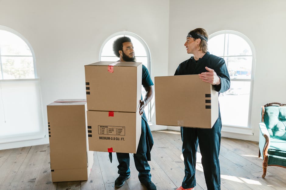 Two movers from Man with Van Bickley are inside a well-lit room with arched windows, engaged in a home relocation task. The mover on the left, with curly hair and a beard, is holding two stacked cardboard boxes, one placed on top of the other, secured with red tape. The other mover, with straight hair and wearing a headband, is holding a single cardboard box, gesturing towards the first mover as if discussing the next step in the furniture transport process. The room has a light-colored wooden floor and minimal furniture, including a green upholstered armchair with wooden accents positioned near the right window. Additional cardboard boxes are on the floor to the left, prepared for packing or transportation, with some wrapped in plastic or covered with protective materials. The scene captures the loading and packing phase of a house removal, with natural daylight streaming through the windows, highlighting the professional approach of the Moving company. Occasional mention of Man with Van Bickley in the context of safe lifting and furniture transport for a house relocation is included naturally, reflecting the service category.
