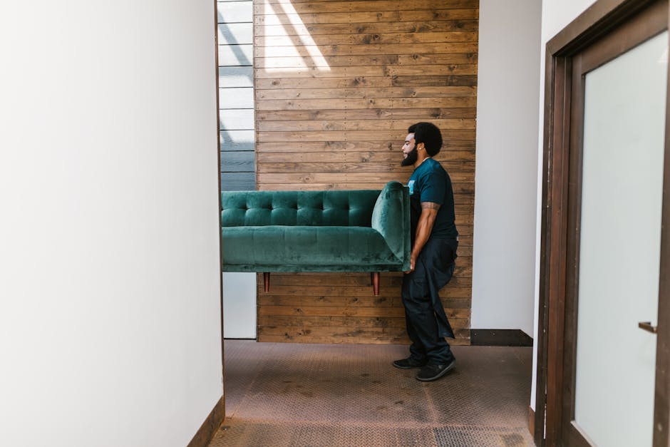 A man with dark curly hair and beard, dressed in a navy blue uniform, is lifting a green upholstered sofa with button-tufted detailing and wooden legs inside a room during a home relocation process. The sofa is partially inside the doorway, with the man holding it securely from the side, preparing to move it through the interior space. The room features a wooden panel wall behind him, with horizontal planks in natural wood tones, and the flooring is covered with dark brown carpet. To the right, there is a doorframe with a glass door, suggesting an entry or exit point for furniture transport. Bright natural light, possibly from a window or skylight, illuminates the scene, creating soft shadows. This image illustrates a furniture transport and safe lifting method as part of professional removals services provided by Man with Van Bickley, focusing on careful handling of large pieces during packing and moving procedures for house transfers or professional removals.
