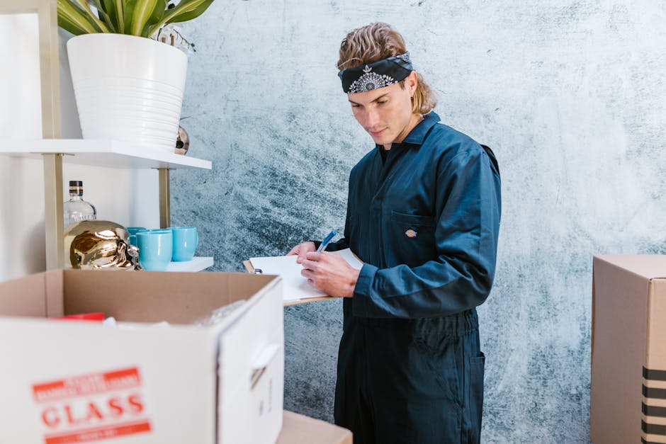 A young man wearing a dark blue jumpsuit and a black bandana with white patterns is inside a room, standing next to a cardboard box labeled 'FRAGILE' on a shelf. He is holding a clipboard and writing with a pen, suggesting he is inspecting or checking items during a home relocation or packing process. Surrounding him are various packing materials, including a large potted plant on a white shelf, porcelain cups and a decorative gold object, all arranged on the shelf. The room has a textured light blue wall and appears to be part of a moving or packing area, with the man focused on documentation related to furniture transport or house removals, aligning with services offered by Man with Van Bickley.