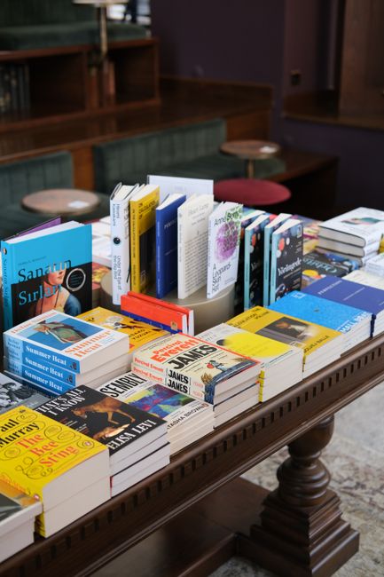 Inside a home library, a woman with short dark hair and light skin, dressed in a light blue shirt and brown trousers, stands near a large bookcase filled with colorful books and cardboard storage boxe