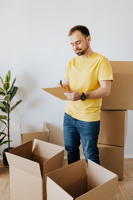 A man in a yellow t-shirt and blue jeans, wearing a black wristwatch, holding a clipboard and pen, standing inside a room surrounded by several open and closed cardboard boxes of various sizes, some with flaps open revealing packing materials. Behind him, there are neatly stacked cardboard boxes ready for a home relocation, with a large indoor plant on the left side of the image, suggesting an organized packing or furniture transport process. The room has white walls and wooden flooring, indicating an interior space prepared for a moving or house removal service. The scene reflects the packing stage of furniture and household items, with visible attention to careful documentation and preparation for client house removals, supporting the services provided by Man with Van Bickley during same-day removals and urgent move logistics.
