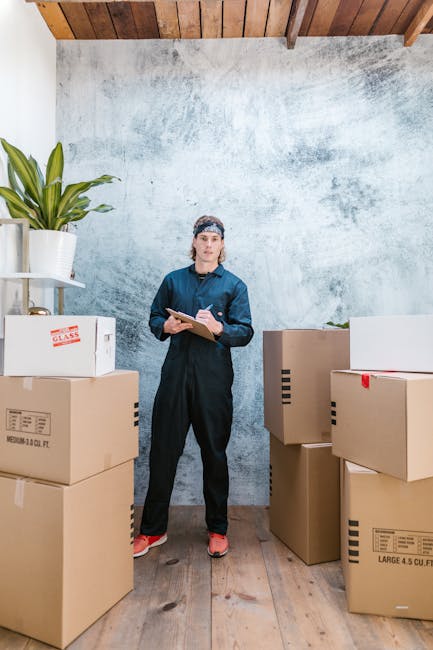 A woman wearing a dark blue uniform and pink shoes stands inside a room with a textured grey wall and a wooden ceiling, surrounded by several cardboard boxes of various sizes, some labeled with weight and size details, and one white flowering plant in a pot placed on a white shelf in the corner. She holds a clipboard and pen, appearing to check or organize packing items as part of her home relocation or furniture transport duties. The boxes are arranged around her on a wooden floor, indicating an ongoing packing or unpacking process related to house removals or moving services provided by Man with Van Bickley. The lighting in the room is natural and bright, emphasizing the workspace for packing and moving logistics.