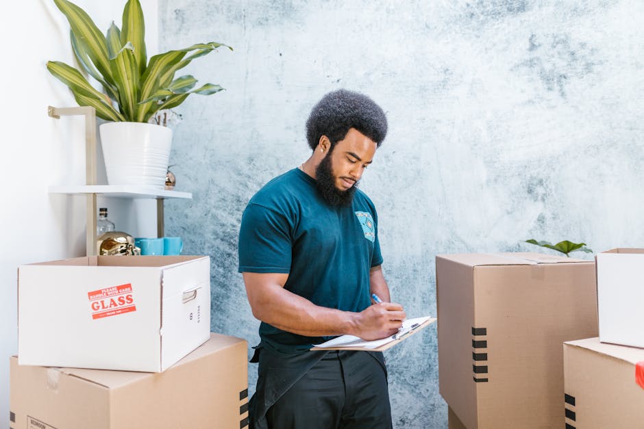 A man in a yellow t-shirt and blue jeans, wearing a black wristwatch, holding a clipboard and pen, standing inside a room surrounded by several open and closed cardboard boxes of various sizes, some with flaps open revealing packing materials. Behind him, there are neatly stacked cardboard boxes ready for a home relocation, with a large indoor plant on the left side of the image, suggesting an organized packing or furniture transport process. The room has white walls and wooden flooring, indicating an interior space prepared for a moving or house removal service. The scene reflects the packing stage of furniture and household items, with visible attention to careful documentation and preparation for client house removals, supporting the services provided by Man with Van Bickley during same-day removals and urgent move logistics.