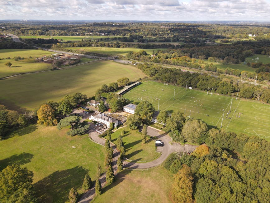 An aerial view of a large residential property set in a rural area, showing a main house with a paved driveway leading to a parking area beside a well-maintained lawn and landscaped garden with tall, narrow trees. Adjacent to the house, there are several cardboard boxes and wrapped furniture, indicating a home relocation process. A manwithvanbickley.co.uk van is parked near the house, with a person loading or unloading large items such as wardrobes, boxes, and furniture from the vehicle onto the driveway area. In the background, there is a sports field with visible goalposts and floodlights, surrounded by abundant greenery, open fields, and distant tree lines under partly cloudy skies. The environment is calm, and the scene captures the logistics of a furniture transport and packing during a local move from Sundridge Park to Bickley, with visible packing materials and the loading process occurring outdoors.