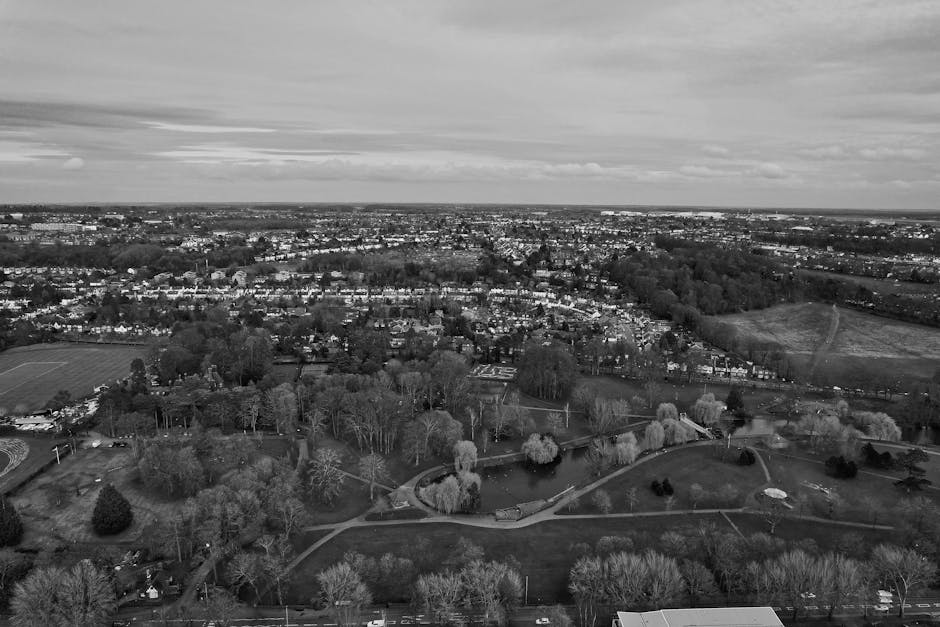 An aerial view of a large residential property set in a rural area, showing a main house with a paved driveway leading to a parking area beside a well-maintained lawn and landscaped garden with tall, narrow trees. Adjacent to the house, there are several cardboard boxes and wrapped furniture, indicating a home relocation process. A manwithvanbickley.co.uk van is parked near the house, with a person loading or unloading large items such as wardrobes, boxes, and furniture from the vehicle onto the driveway area. In the background, there is a sports field with visible goalposts and floodlights, surrounded by abundant greenery, open fields, and distant tree lines under partly cloudy skies. The environment is calm, and the scene captures the logistics of a furniture transport and packing during a local move from Sundridge Park to Bickley, with visible packing materials and the loading process occurring outdoors.